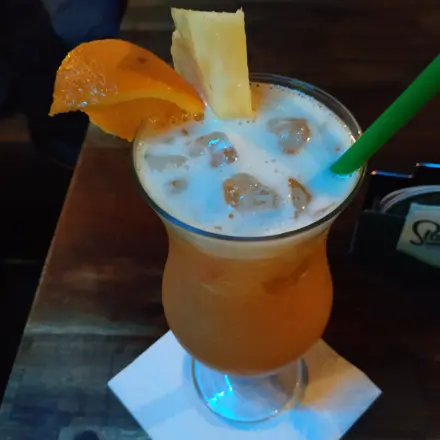 A tall orange cocktail with pineapple and a green straw